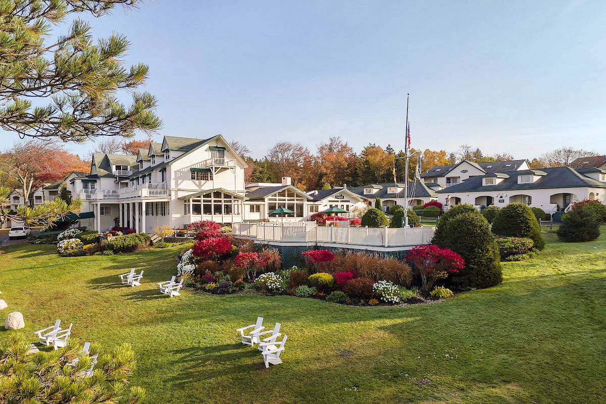 A landscaped garden with colorful flowers, a flagpole, and white buildings in the background, along with white chairs on a well-maintained lawn.