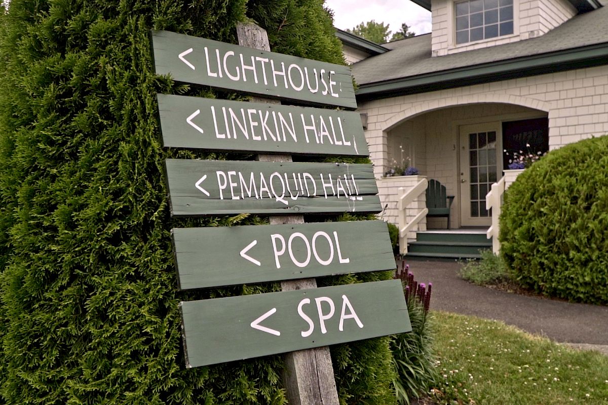A signpost is in front of a building, directing to the lighthouse, Linekin Hall, Pemaquid Hall, pool, and spa, surrounded by greenery.