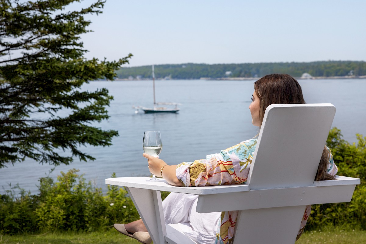 A person sits in a white Adirondack chair, holding a wine glass, and looks out at a scenic lake with a boat in the distance and trees on the shore.