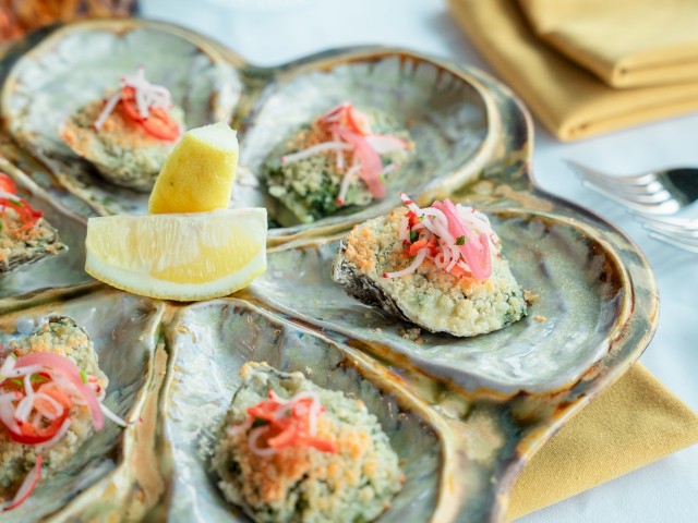 A beautifully plated seafood dish with six shells, each topped with breadcrumb mixture and garnish, and lemon wedges in the center.