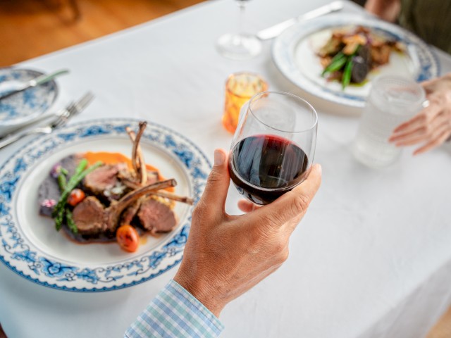 Two people dining with meals on decorative plates; one holds a glass of red wine, the other a glass of water; elegant table setting.