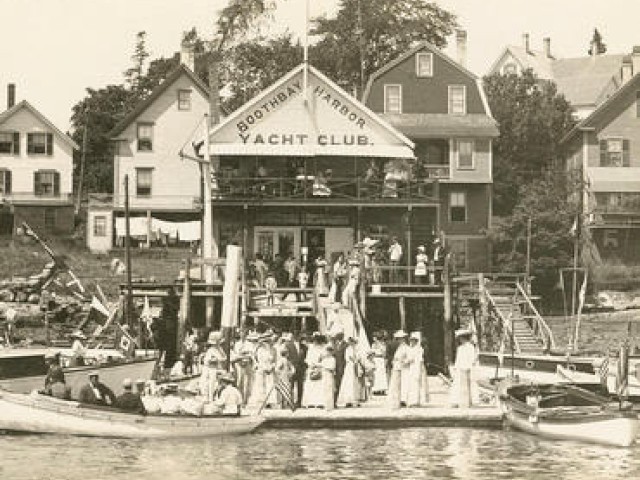 A group of people are gathered in front of the Boothbay Harbor Yacht Club, with several boats docked by the waterside, ending the sentence.