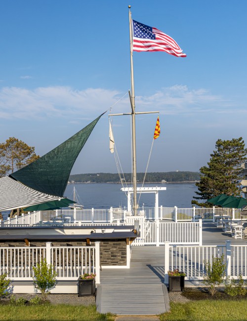 A deck with a flagpole, an American flag, and a scenic view of water and trees in the background, surrounded by a white fence, is pictured.