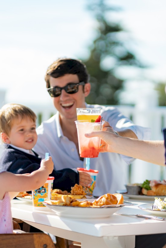 A family enjoys a meal at an outdoor table, with two adults and two children sharing food and drinks, smiling and toasting each other.