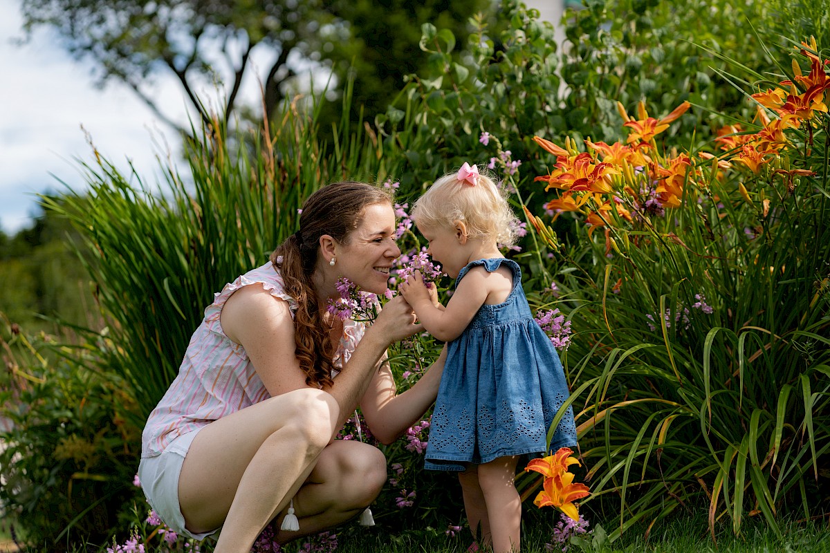 A woman and a child are smelling flowers in a garden, surrounded by lush greenery and vibrant orange lilies.