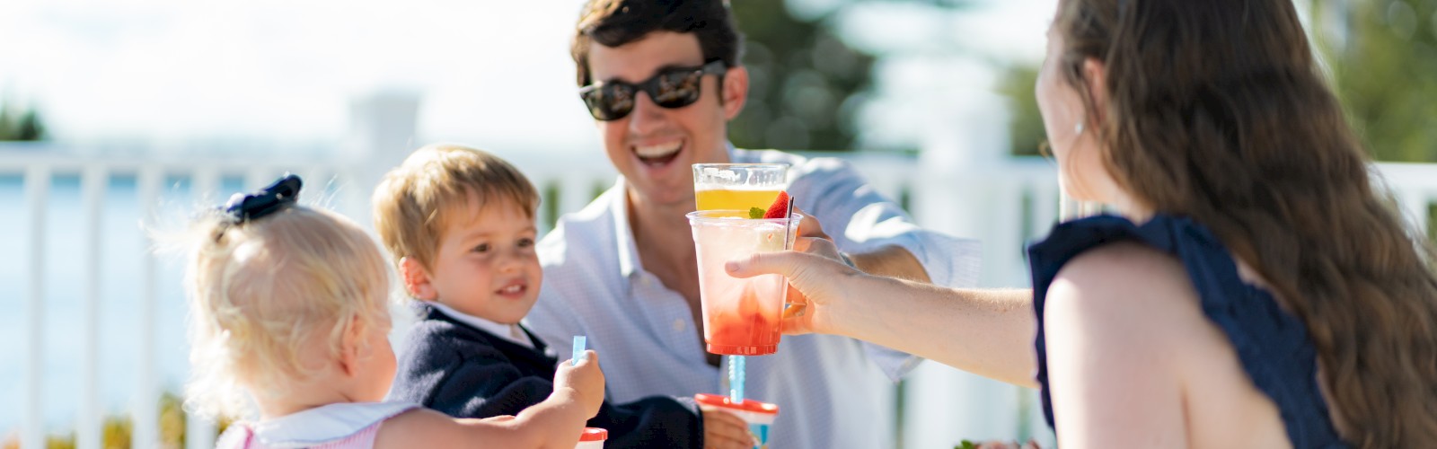 A family enjoys an outdoor meal together; two adults toast drinks while two children are at the table with food, creating a cheerful scene.