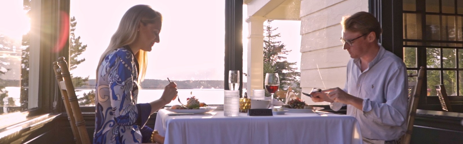 A couple is dining together at a table near large windows with a scenic view of water and trees during sunset.