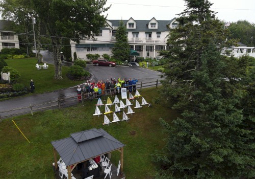 A group of people standing near small tents in front of a large house, surrounded by trees and a gazebo.