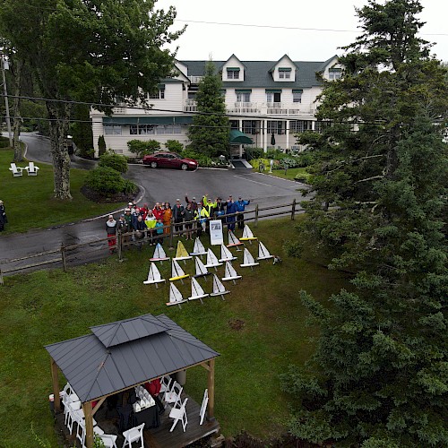 A group of people standing near small tents in front of a large house, surrounded by trees and a gazebo.