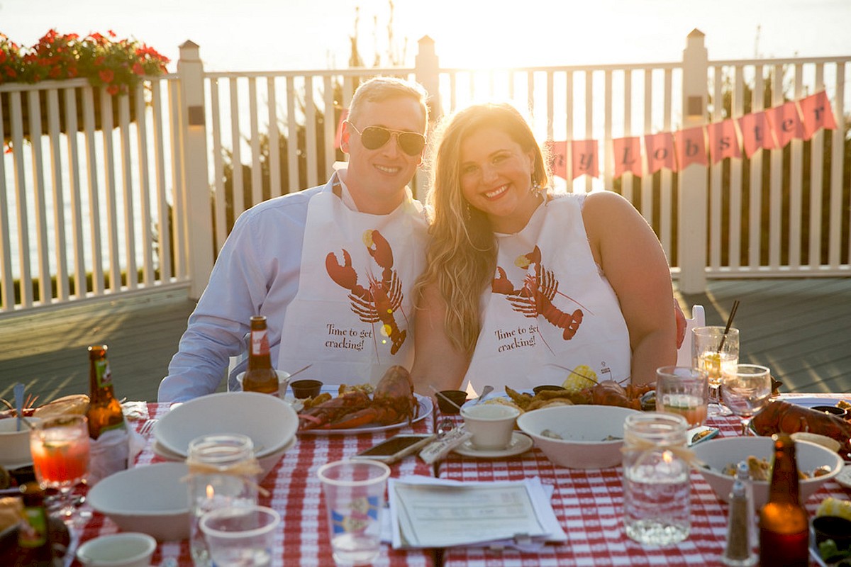 Two people wearing lobster bibs sit at a table with food and drinks on a checkered tablecloth, while sitting together.