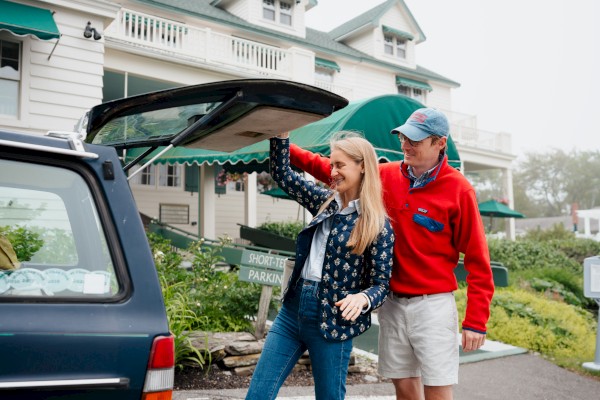 A couple loads a trunk at a roadside event; woman in a blue plaid shirt and jeans helps as a man in a red sweater and cap assists, near a white house and green awnings.