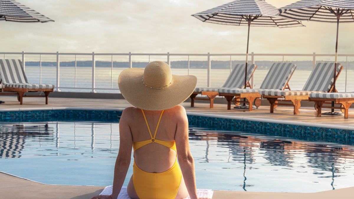 A person in a yellow swimsuit sits by a pool, wearing a wide-brimmed hat. Lounge chairs and striped umbrellas are in the background.