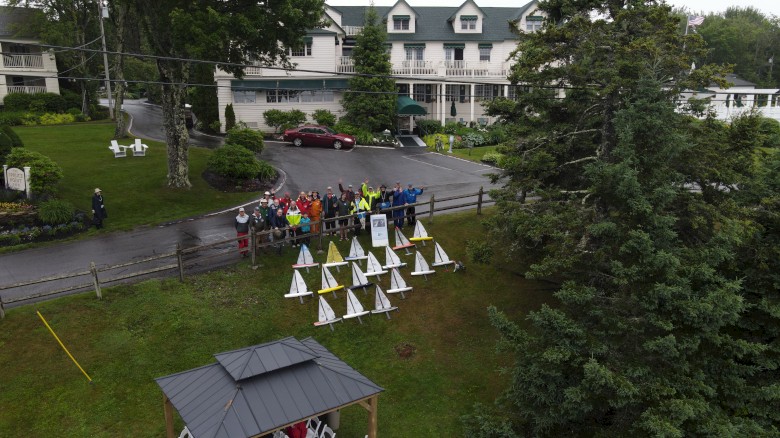 A group of people stands near triangular structures outdoors, with a white building and trees in the background.