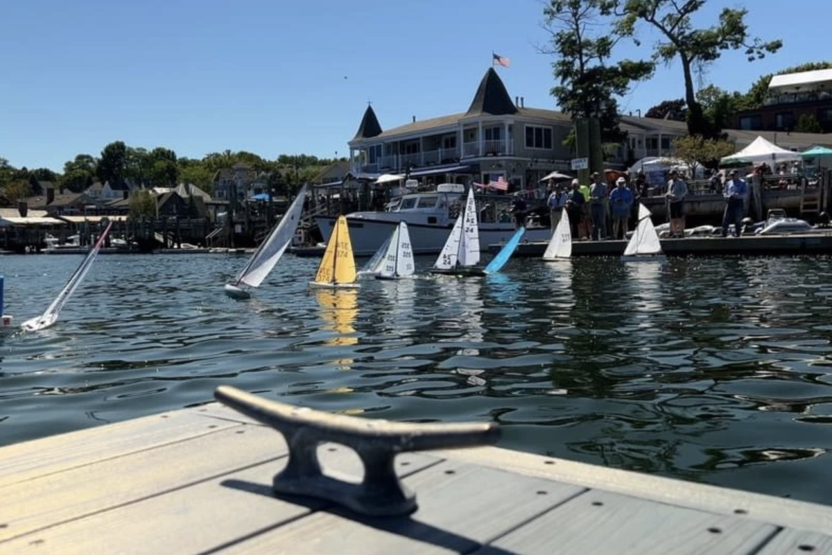 Model sailboats on the water near a dock, with a dock cleat in the foreground, and a waterfront building and spectators in the background.