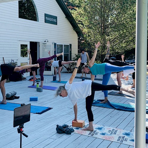 People are practicing yoga on a deck outside a building, using mats and blocks, with a camera on a tripod recording the session.
