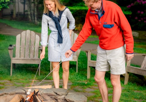 Two people are toasting marshmallows over a fire pit in a garden setting, surrounded by trees and chairs.