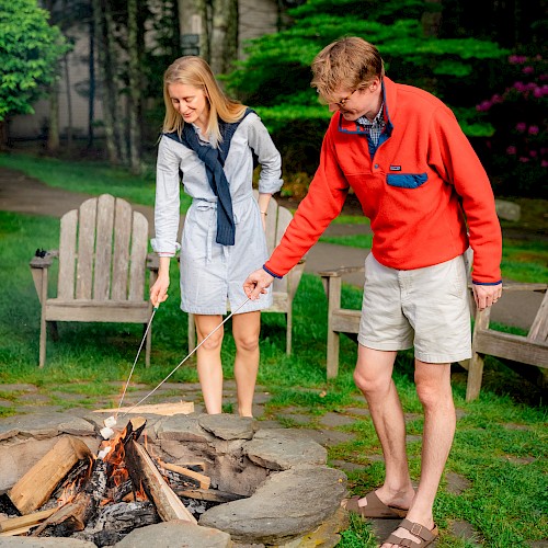 Two people are toasting marshmallows over a fire pit in a garden setting, surrounded by trees and chairs.