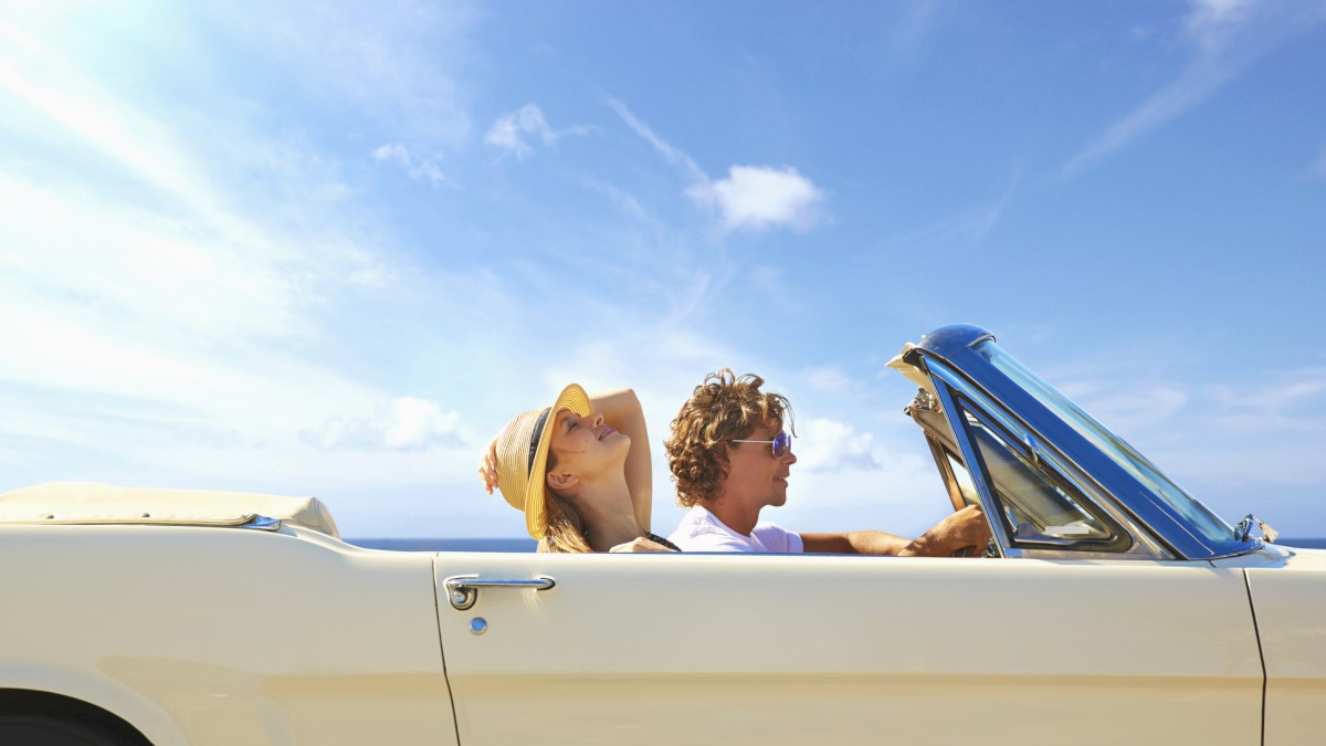 Two people are enjoying a sunny day in a convertible car, with the top down and blue skies overhead.