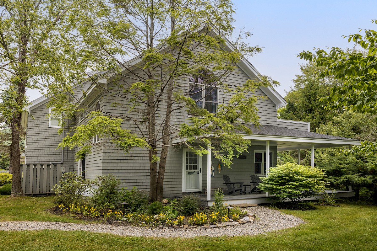 A charming gray house with a well-maintained garden, a gravel path, and a covered porch surrounded by trees and greenery.