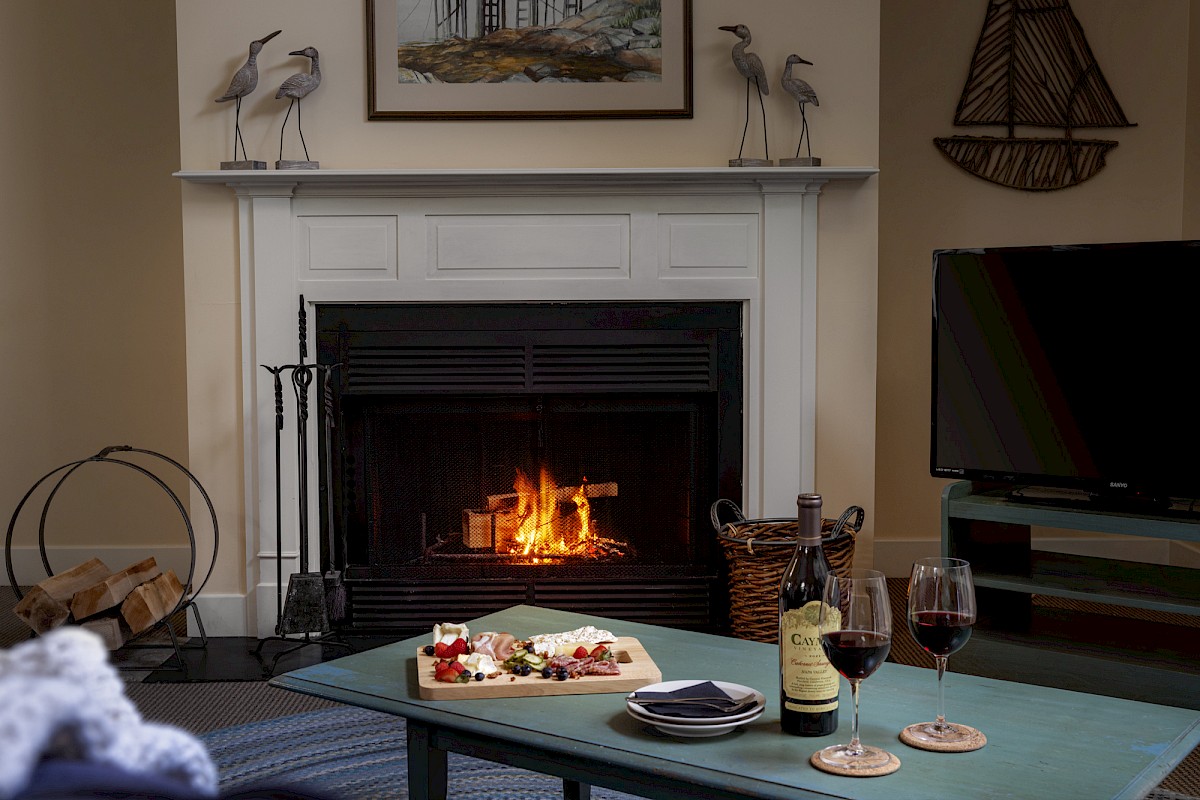 A cozy living room with a lit fireplace, a coffee table with red wine and snacks, and a TV on a stand.