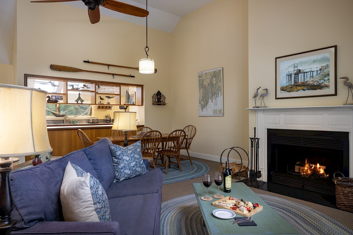 A cozy living room with a blue sofa, a fireplace, a coffee table with food and wine, and a dining area near the kitchen under a high ceiling.