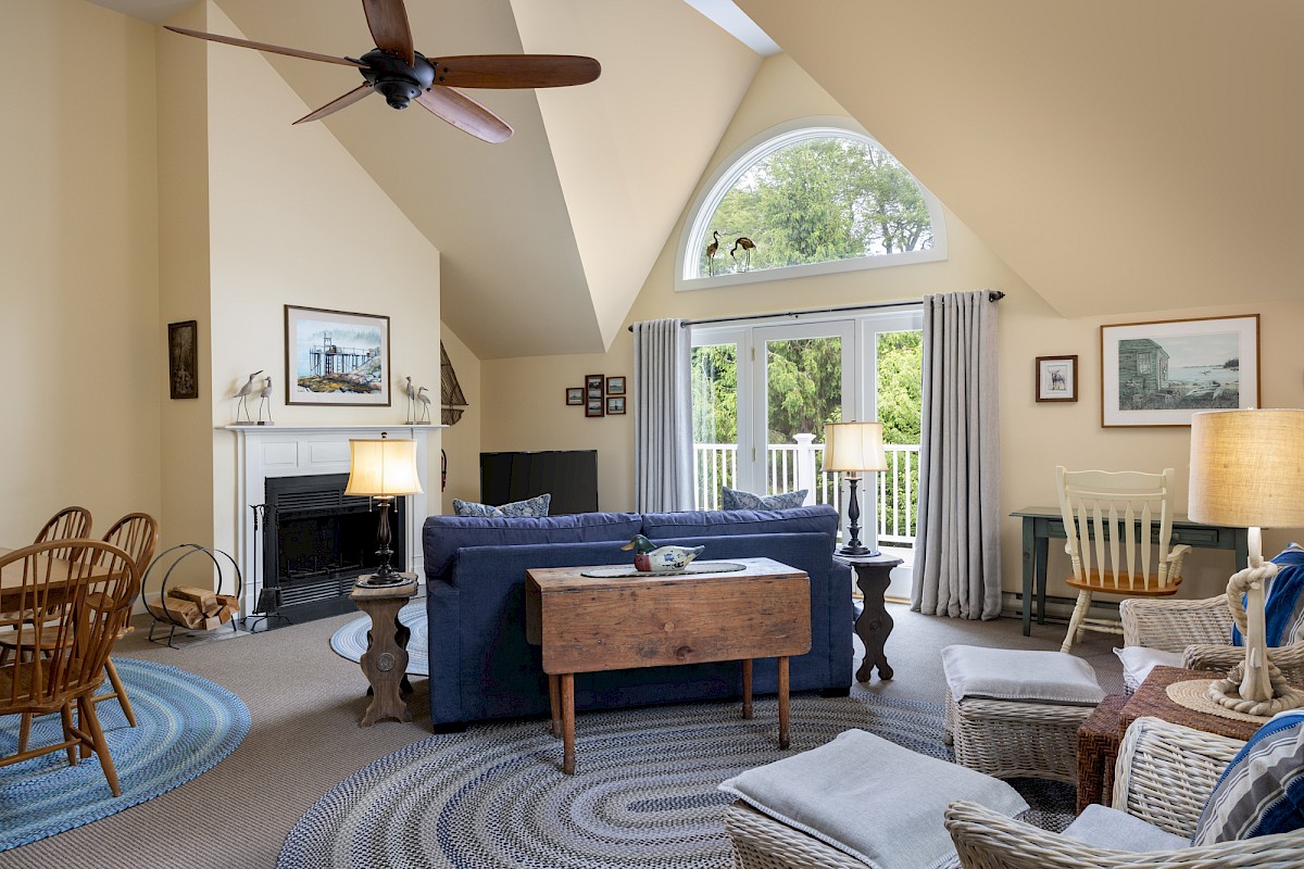 A cozy living room with a vaulted ceiling, blue sofa, wicker chairs, and a wooden coffee table. Large window with drapes and framed art.
