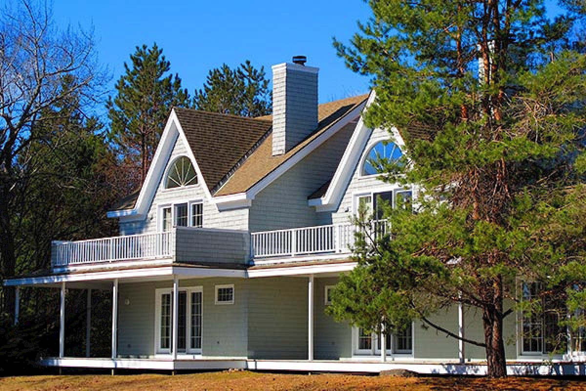 The image shows a gray two-story house with a unique roof design, two chimneys, a large balcony, and surrounded by trees and a clear blue sky.