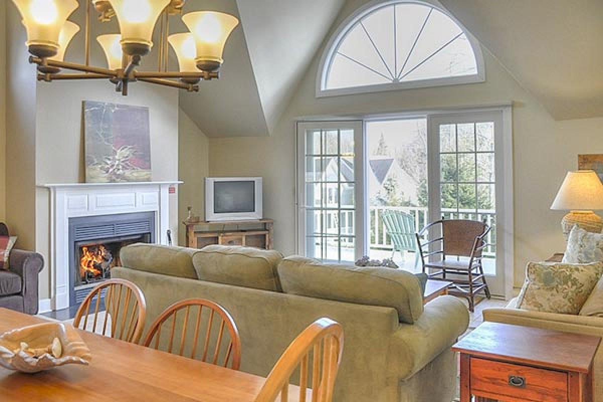 A cozy living room with a fireplace, TV, large windows, and a chandelier above the dining table. There is seating for multiple people.
