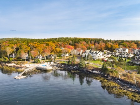 A scenic coastal village with houses, a dock, and a tree-lined background showing fall foliage by the water.