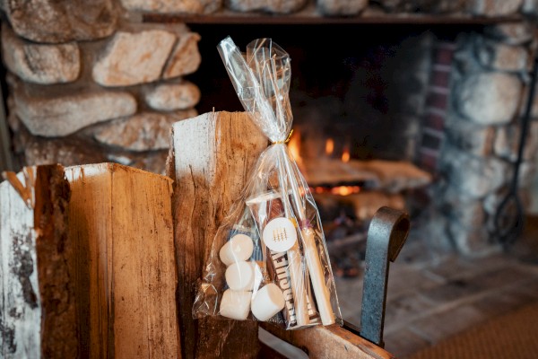 A cozy fireplace with a bag of marshmallows and chocolates, suggesting s'mores, next to stacked firewood.