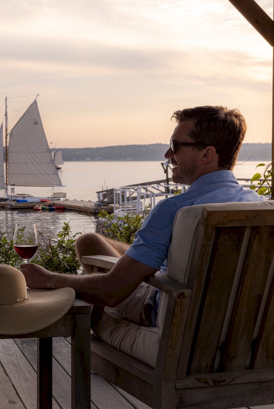 Two people sit on a deck with wine, watching a sailboat on the water at sunset. A hat and a wine bottle are on the table between them.