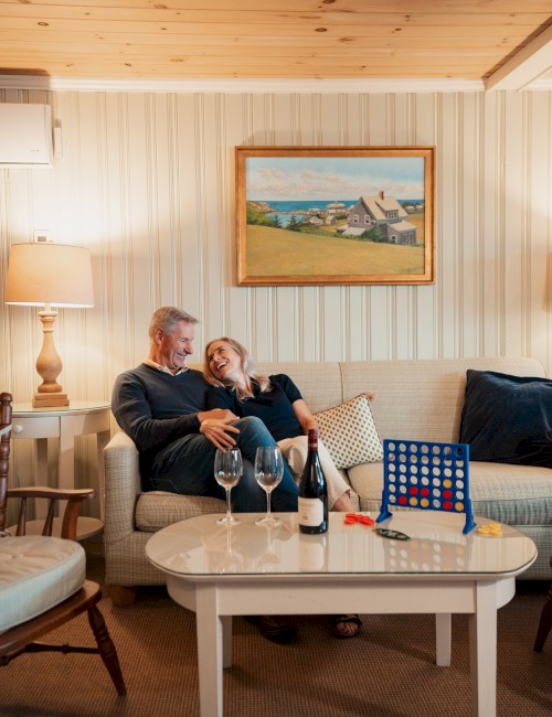 A couple relaxes on a sofa, surrounded by wine glasses and a Connect Four game, set in a cozy room with a lamp and landscape painting.