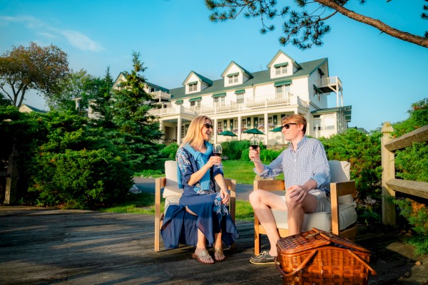 Two people sit chatting on a wooden deck in front of a large white multi-story house with green trees around, enjoying a sunny day on a picnic.