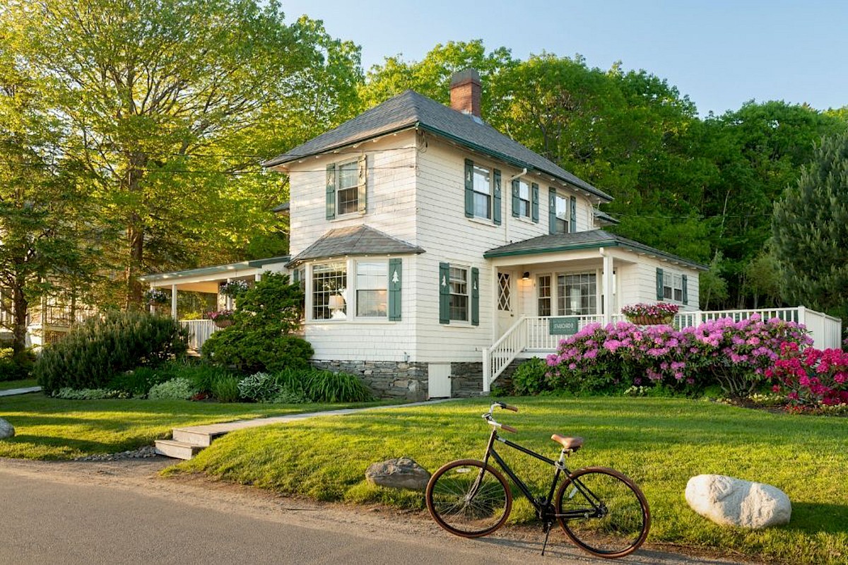 A charming two-story house with a porch and garden, surrounded by trees and flowers. A bicycle is parked on the grass nearby.