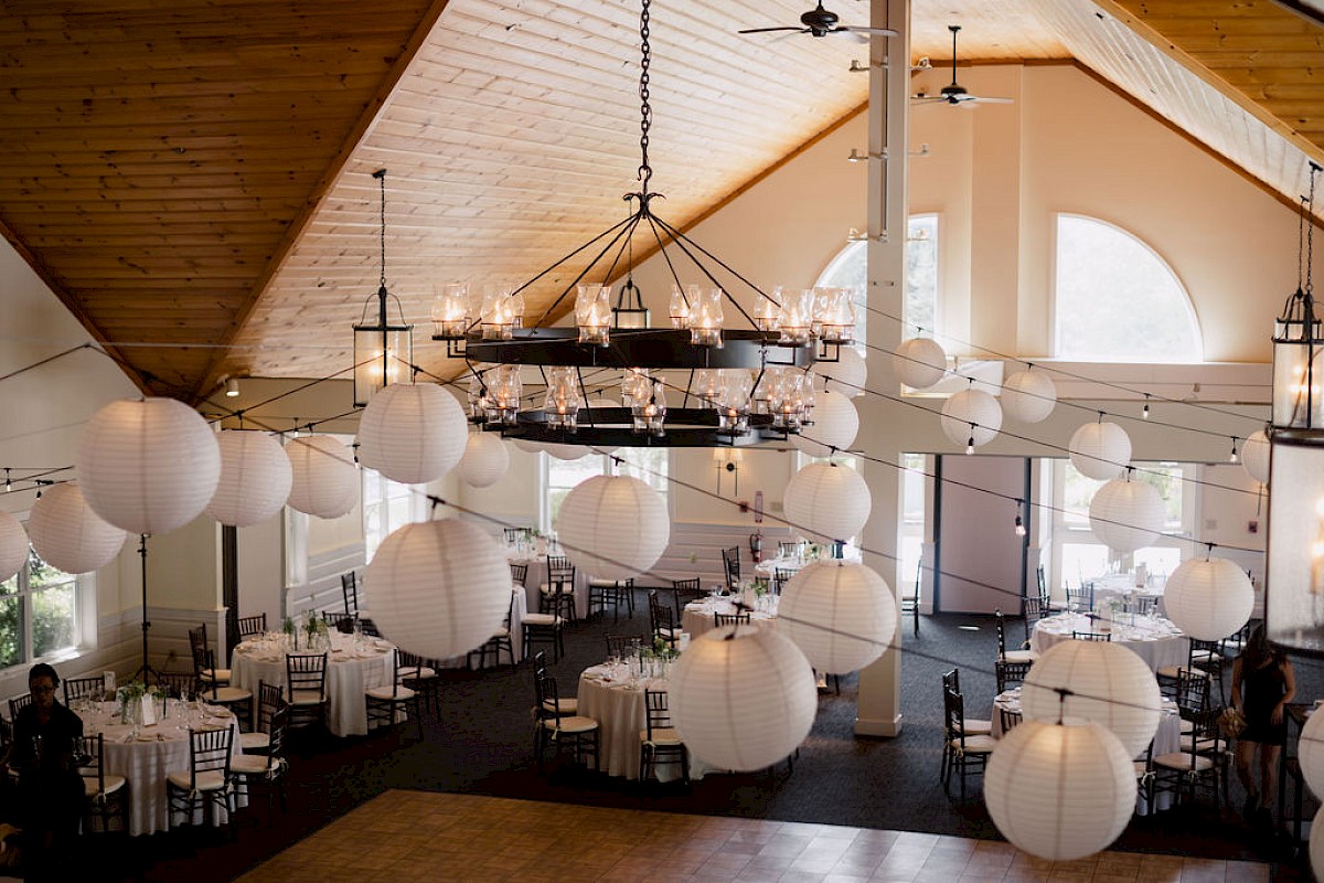 A decorated indoor event space with tables, chairs, chandeliers, and large hanging lanterns, creating a festive atmosphere.