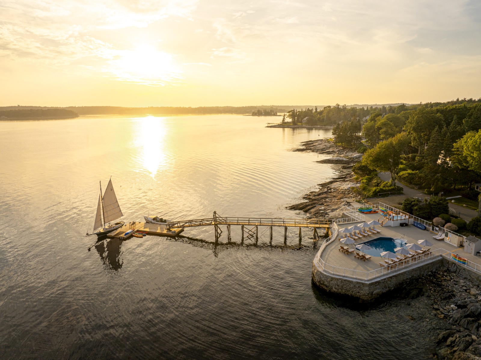 A scenic coastal view at sunrise with a sailboat at a dock, a pool area with umbrellas, and lush greenery along the rocky shoreline.