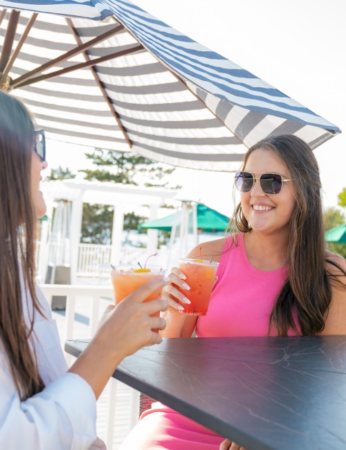 Two people sitting outdoors under umbrellas, enjoying drinks and smiling at each other, capturing a joyful moment in the sun.