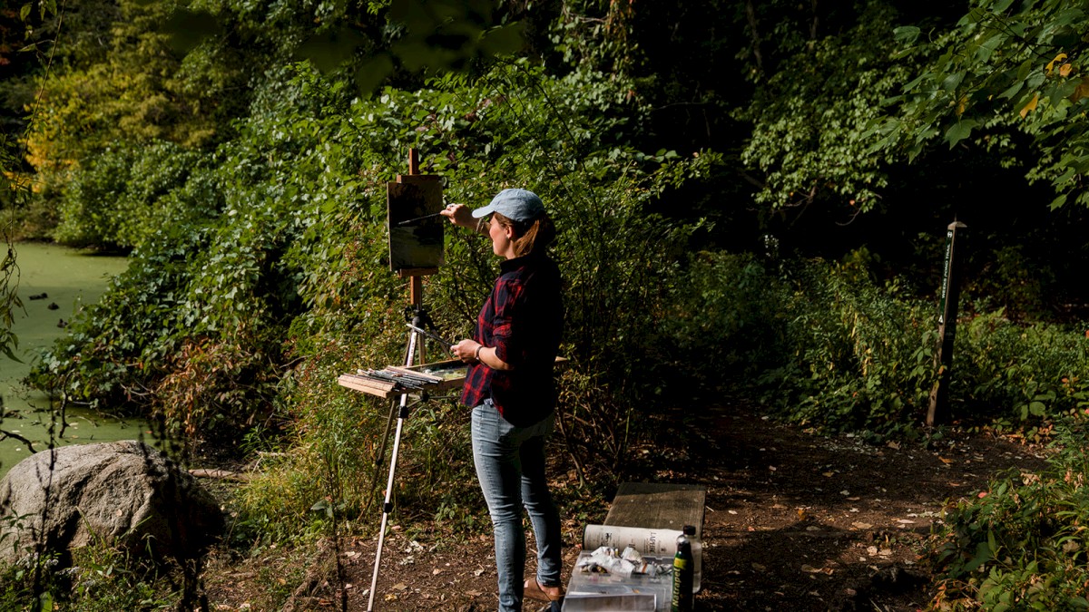 A person paints on an easel in a forest, surrounded by lush greenery and a pond, wearing casual attire and a cap.