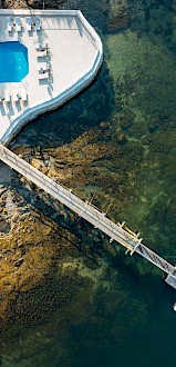 Aerial view of a poolside area with loungers, adjacent to a rocky shoreline, connected by a dock with small boats in the water below.