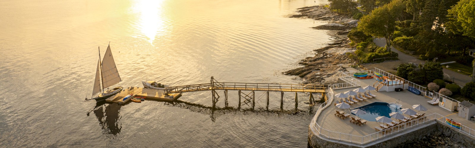 A tranquil coastal scene with a dock, sailboat, and an ocean-side pool on a rocky shore during sunset, surrounded by lush greenery.
