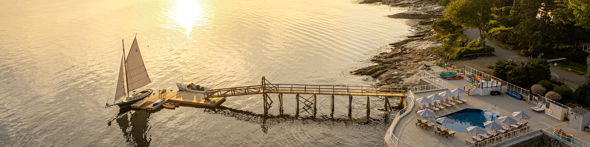 A tranquil coastal scene with a dock, sailboat, and an ocean-side pool on a rocky shore during sunset, surrounded by lush greenery.