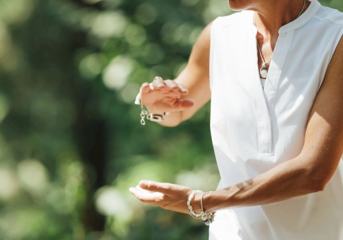 A person gesturing outdoors, wearing a sleeveless white top with bracelets, set against a blurred green background.