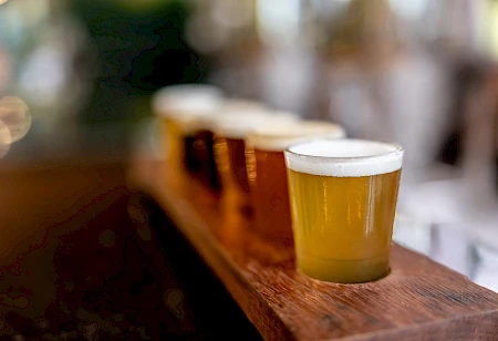A flight of different beers in small glasses on a wooden tray, likely in a pub or brewery setting.