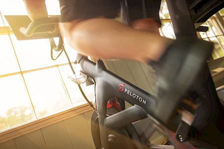A person is exercising on a Peloton stationary bike indoors, with sunlight streaming through the window.