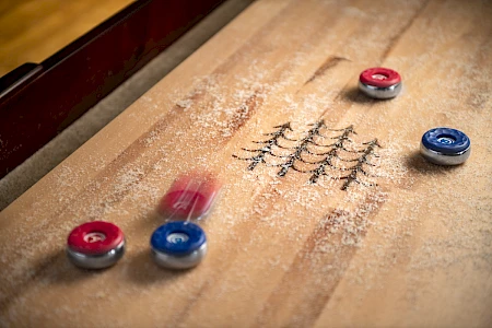 A tabletop shuffleboard game in action, with red and blue pucks sliding on a wooden surface dusted with powder.