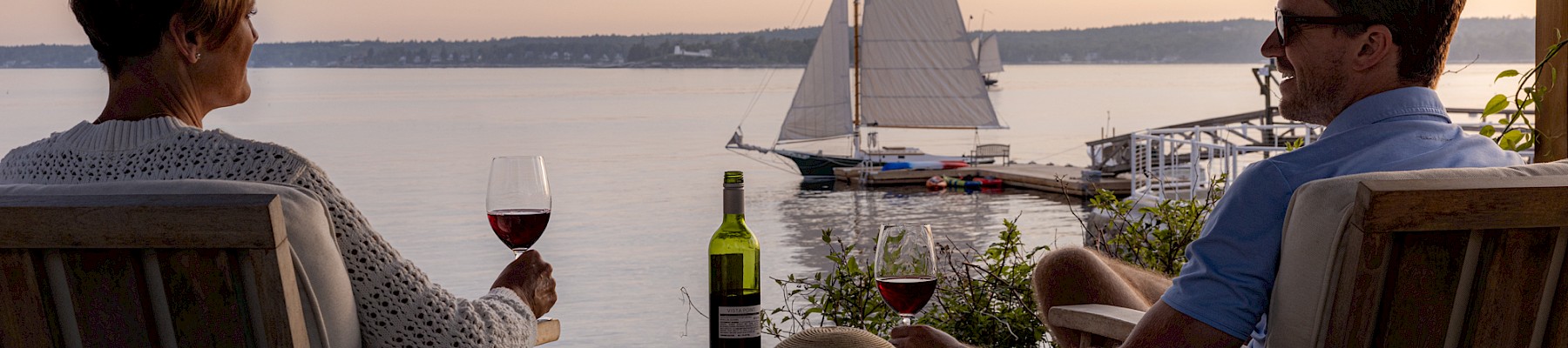 Two people relax on a deck with wine, enjoying a view of a sailboat on the water at sunset.