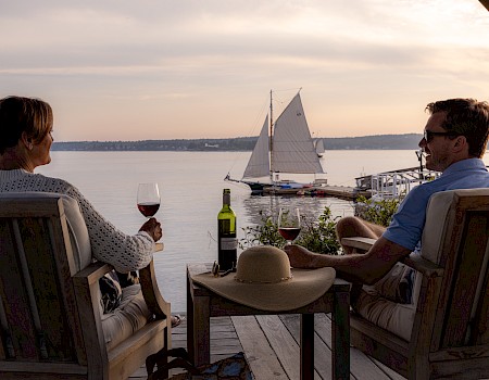 Two people relax on a deck with wine, enjoying a view of a sailboat on the water at sunset.