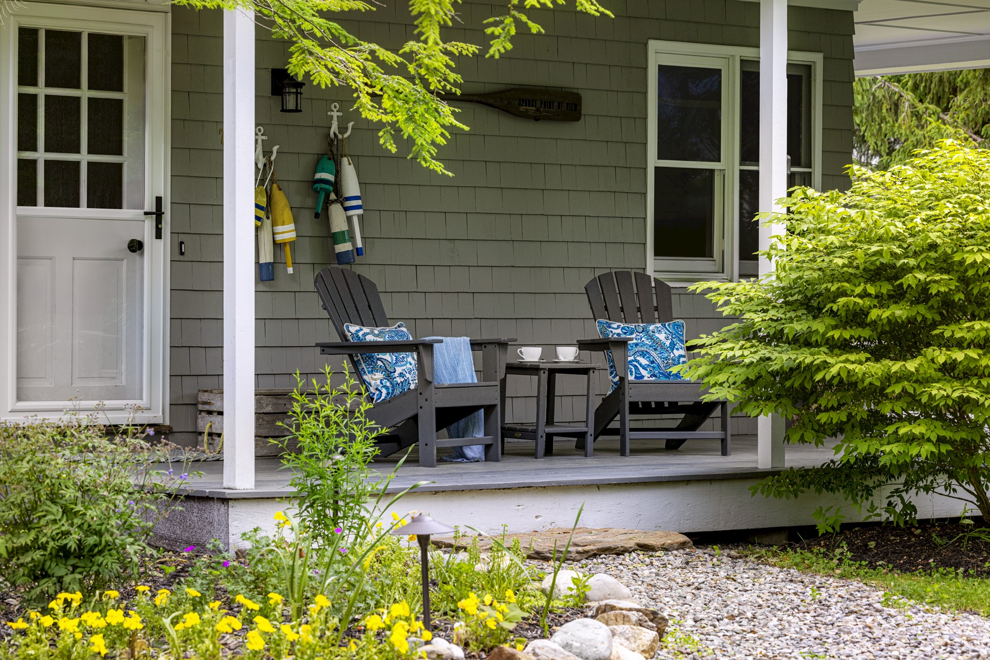 A cozy porch with two Adirondack chairs, colorful pillows, and a small table, surrounded by lush greenery and flowers in bloom.