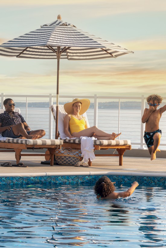 A poolside scene with two people lounging under umbrellas, a child jumping into the pool, and another swimming, against a sunset backdrop.