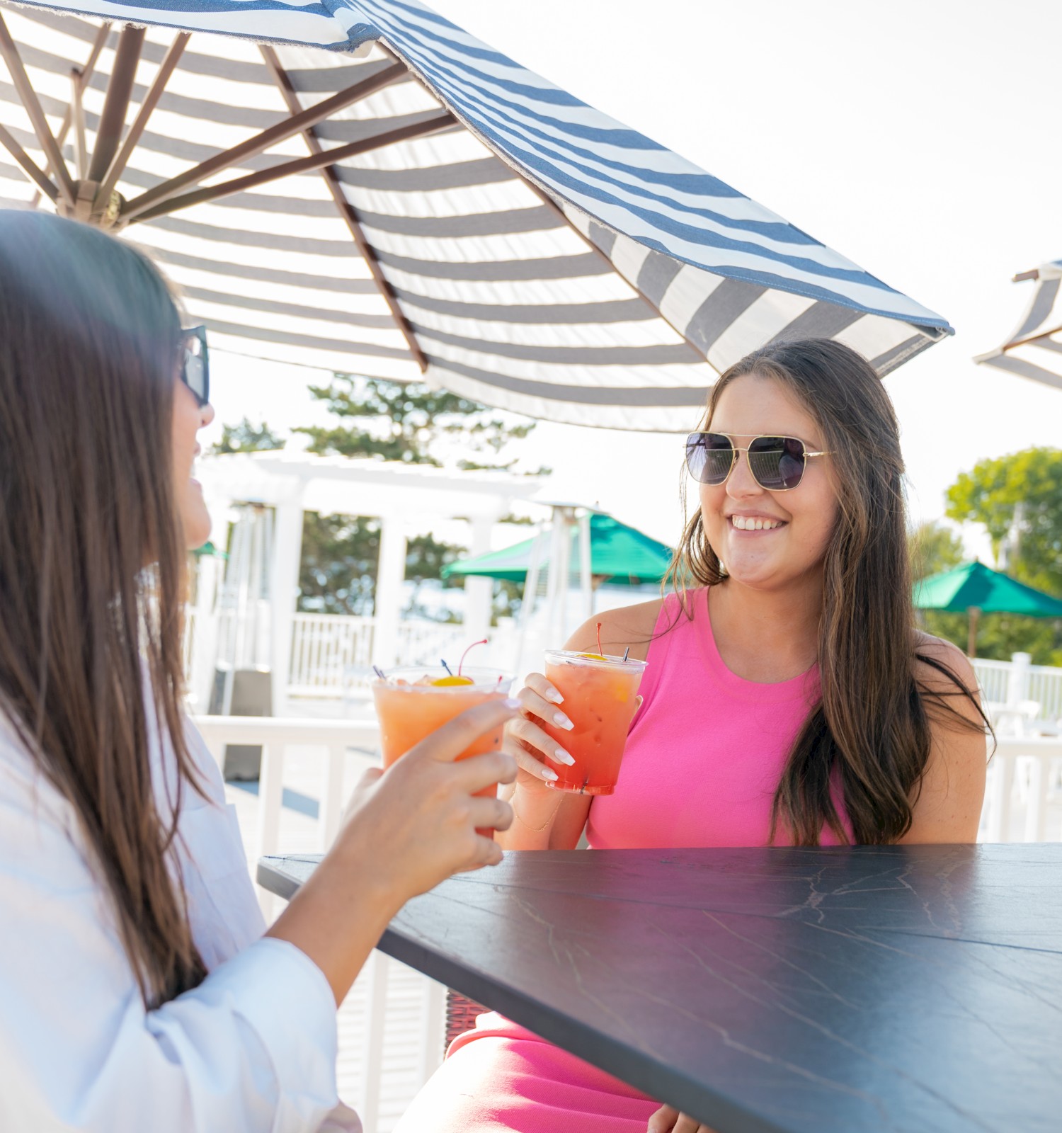 Two people sitting outdoors under umbrellas, enjoying drinks and each other's company, with a bright and sunny atmosphere.